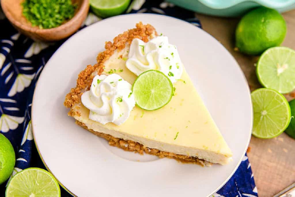 A slice of key lime pie with a graham cracker crust, topped with whipped cream and a lime slice, served on a white plate with fresh limes nearby.