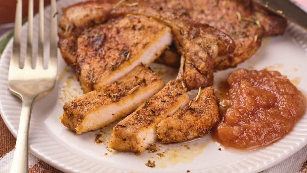 A plate with seasoned, sliced pork chop and a serving of applesauce next to a fork.