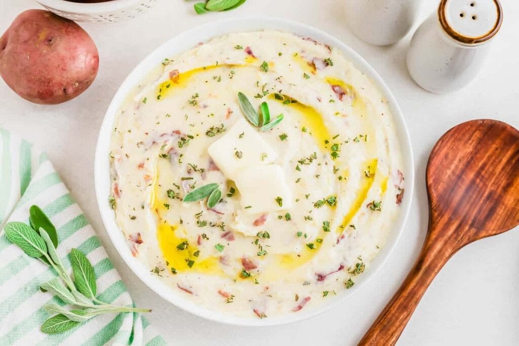 Bowl of mashed potatoes with herbs, a pat of butter, and a drizzle of oil, next to a wooden spoon, salt and pepper shakers, a red potato, and fresh sage leaves.