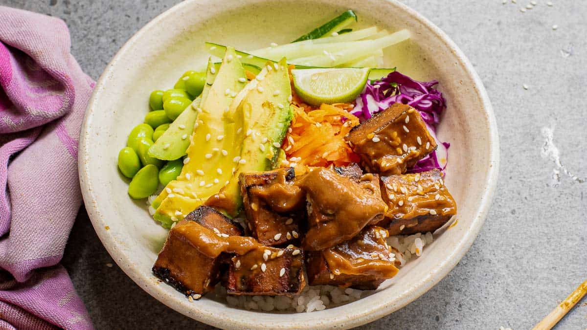 Bowl with rice, tofu cubes, avocado slices, edamame, cucumber, red cabbage, carrot, and lime, topped with sesame seeds and sauce. Pink cloth on the side.