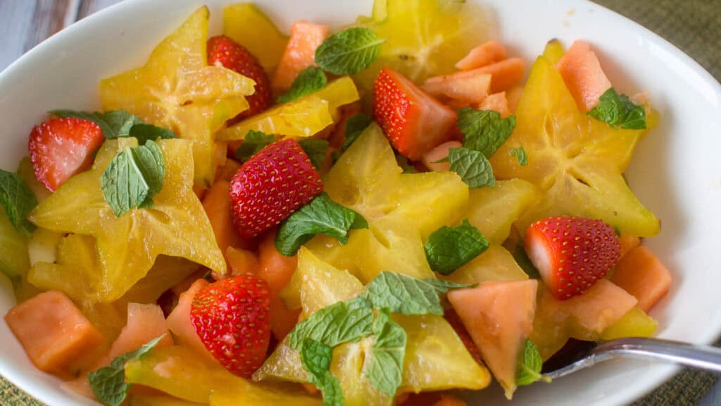 A white bowl filled with a fruit salad featuring starfruit slices, strawberries, papaya chunks, and mint leaves.
