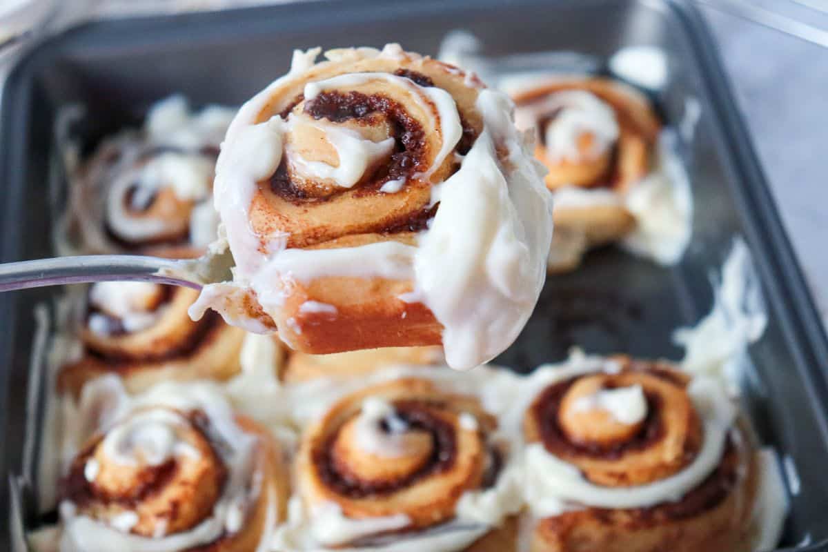 A close-up of a cinnamon roll with white icing being lifted from a pan filled with more iced cinnamon rolls.
