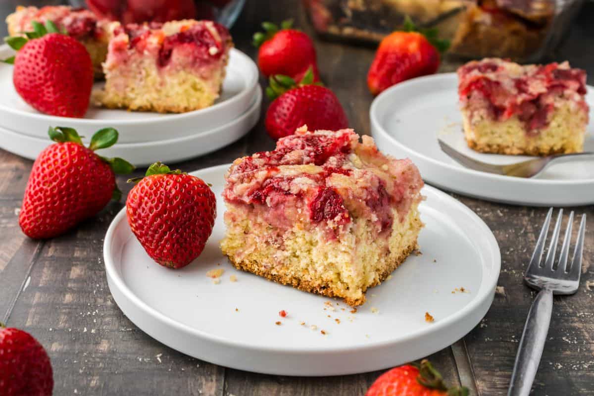 A slice of strawberry crumb cake on a white plate, surrounded by fresh strawberries, with more slices on plates in the background.