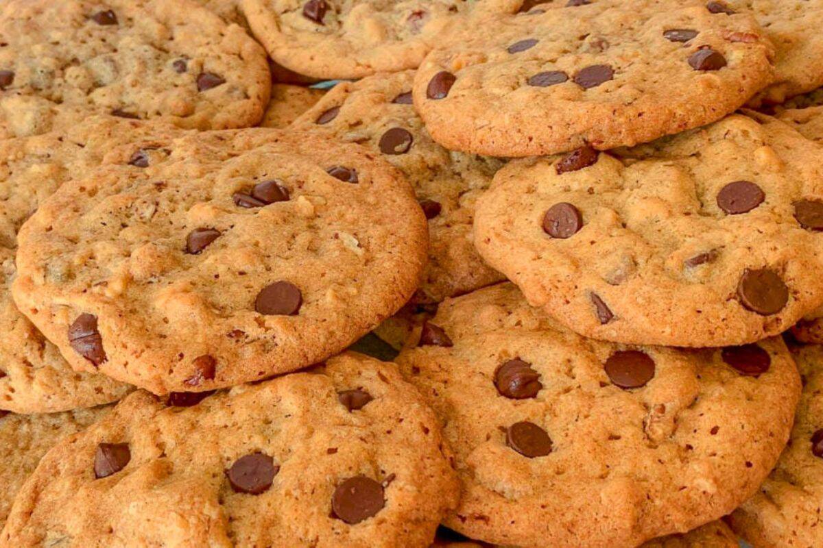 A close-up of a pile of chocolate chip cookies with golden-brown, crispy edges and scattered chocolate chips on top. The cookies have a textured surface and appear freshly baked.