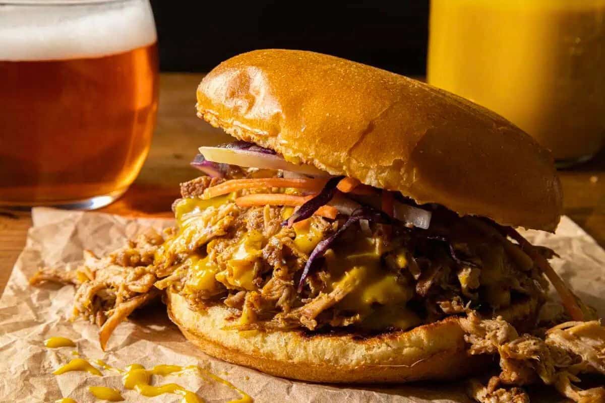 A delicious showcase of Southern cooking, this pulled pork sandwich with melted cheese, pickles, and shredded veggies is served on a bun with brown paper—perfect alongside a glass of beer and a yellow drink in the background.