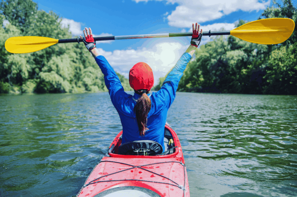Person in a red kayak raises a yellow paddle overhead, enjoying the calm river and green trees under a partly cloudy sky&mdash;an adventure often found at adult summer camps.