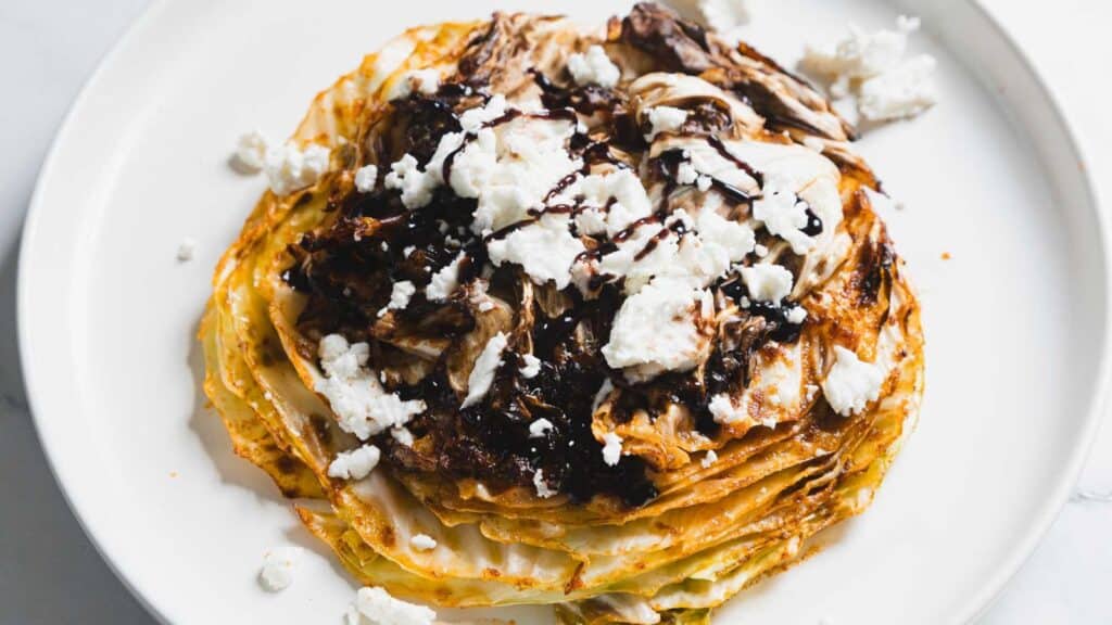 A stack of roasted cabbage slices topped with crumbled cheese and a drizzle of dark sauce on a white plate.