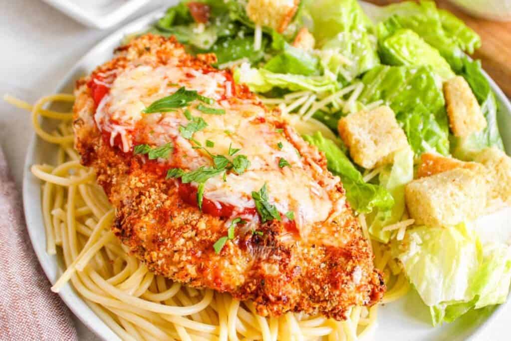 A plate with breaded chicken topped with tomato sauce and cheese on spaghetti, served with a green salad and croutons.