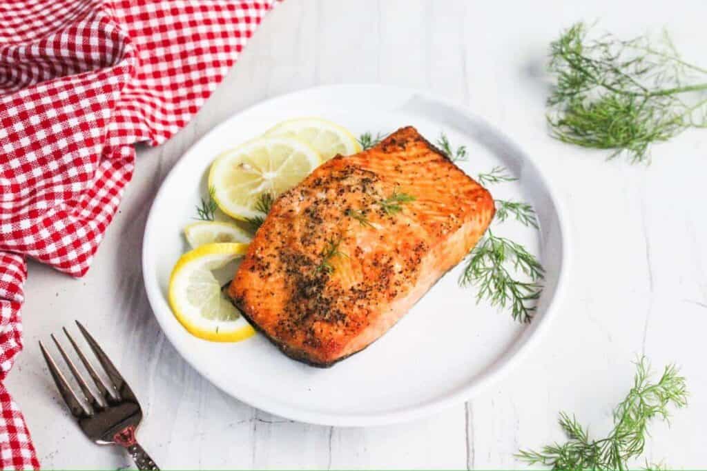 A cooked salmon fillet garnished with dill rests on a white plate with lemon slices, next to a fork and a red checkered cloth.