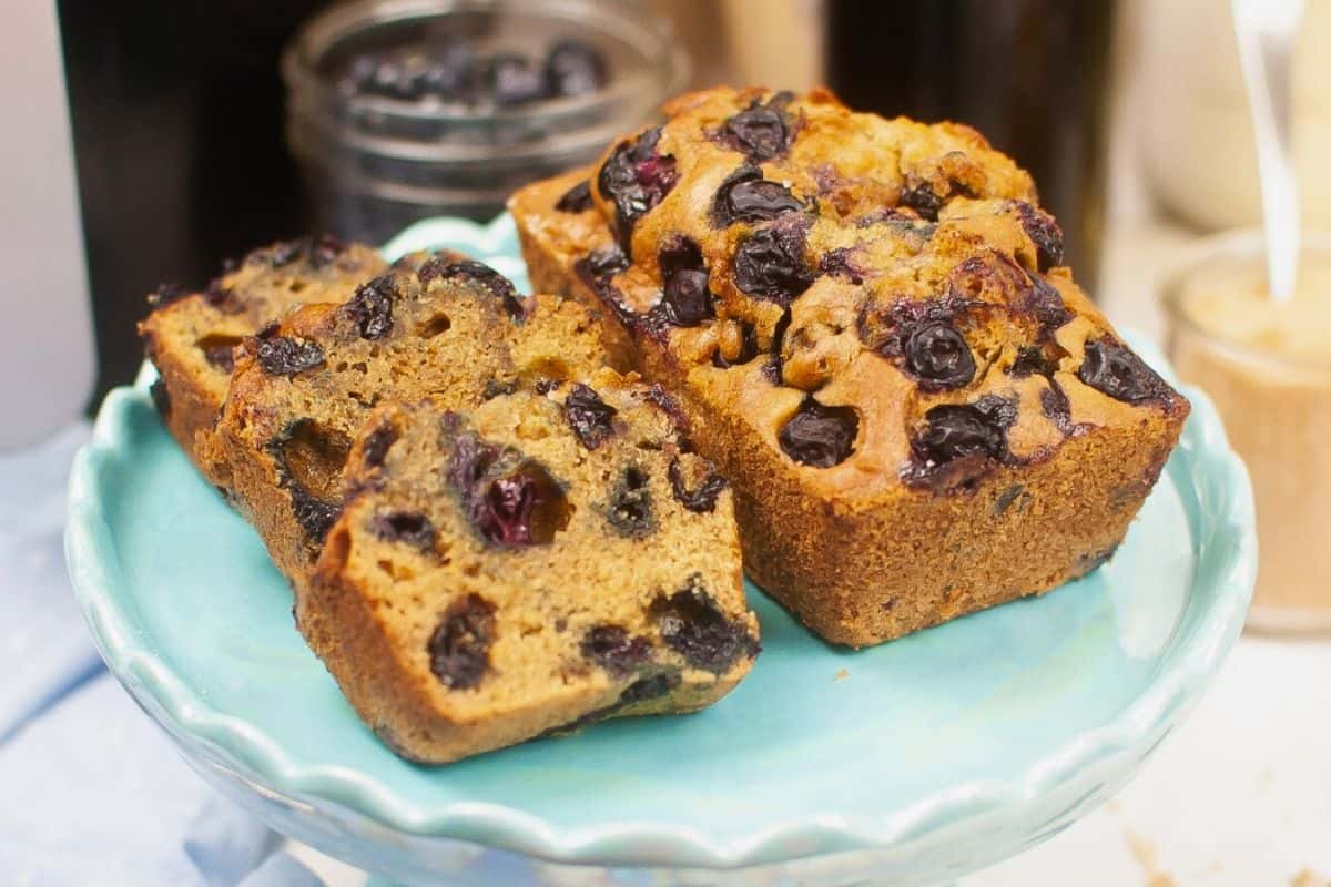 Two slices of blueberry bread on a light blue plate, with a whole loaf behind them and a jar of blueberries in the background.
