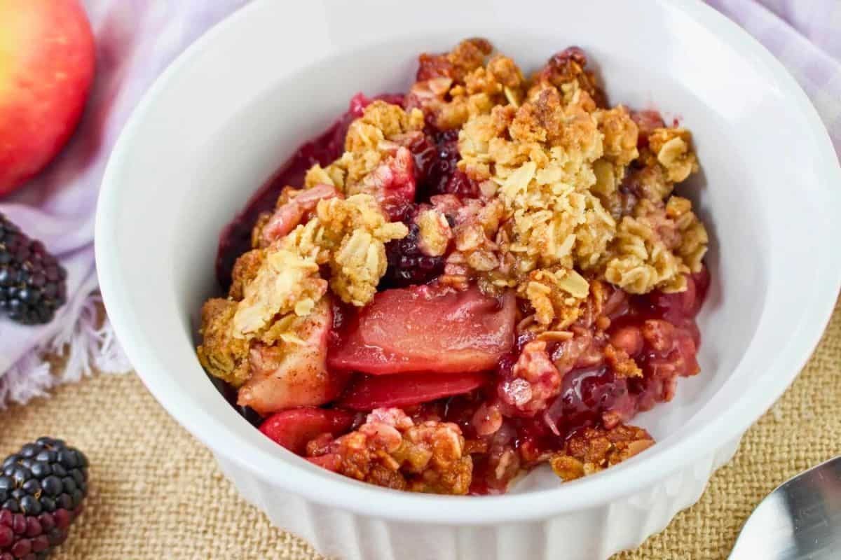 A bowl of fruit crumble with oats on top, featuring apples and blackberries. A fresh apple and blackberries are placed beside the bowl.