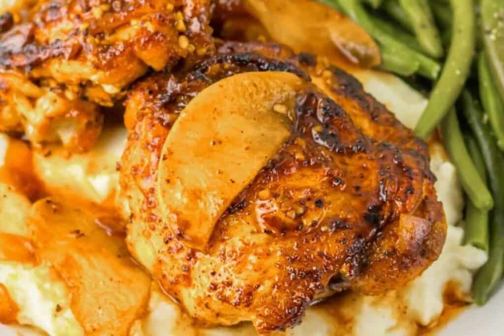 A cooked chicken thigh with apple slices on mashed potatoes, served with green beans.