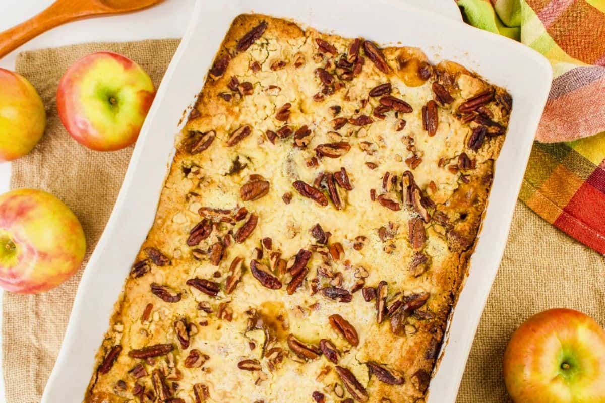 A baked apple dessert topped with pecans in a rectangular dish, surrounded by whole apples and a colorful napkin.