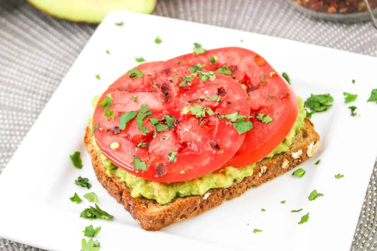 A slice of whole-grain bread with avocado spread topped with two tomato slices and garnished with chopped cilantro on a white square plate.