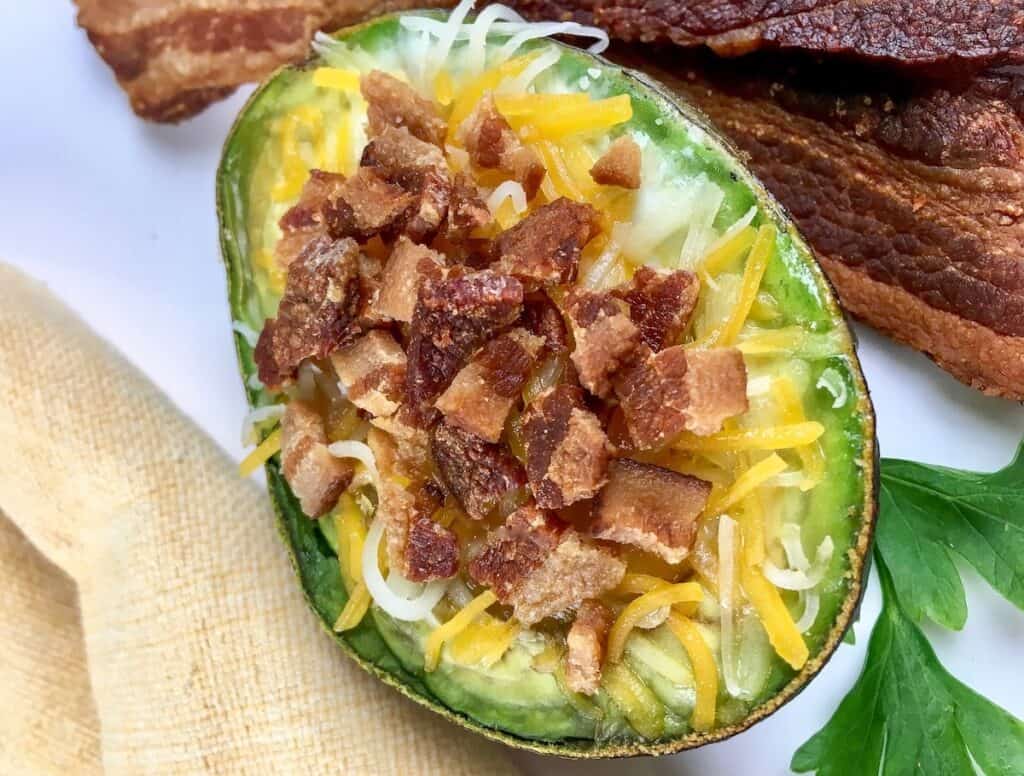 Half an avocado filled with shredded cheese and topped with chopped bacon, placed on a plate next to bacon strips and a sprig of parsley.