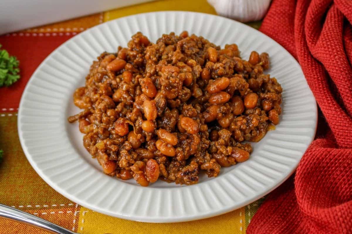 A plate of baked beans mixed with ground beef, placed on a white plate. The plate is on a colorful tablecloth, with a red cloth and greens nearby.