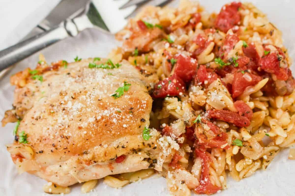 A plate with baked chicken thigh and orzo pasta mixed with tomatoes, garnished with herbs, with a fork and knife beside.