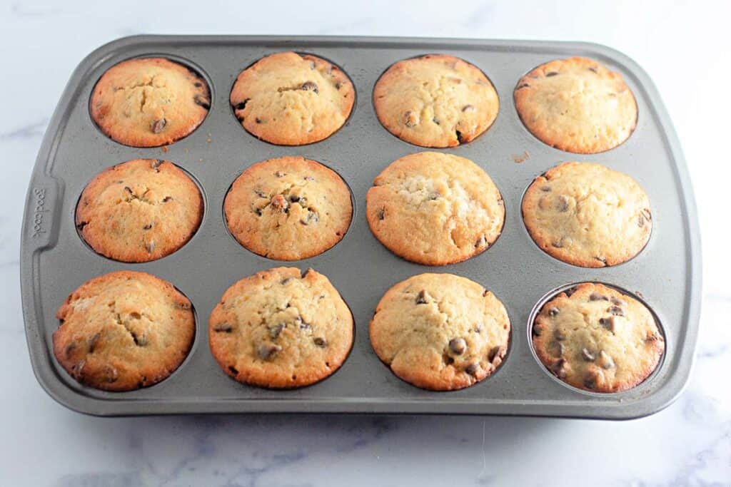 A metal muffin tray containing twelve baked chocolate chip muffins, viewed from above on a light-colored surface.