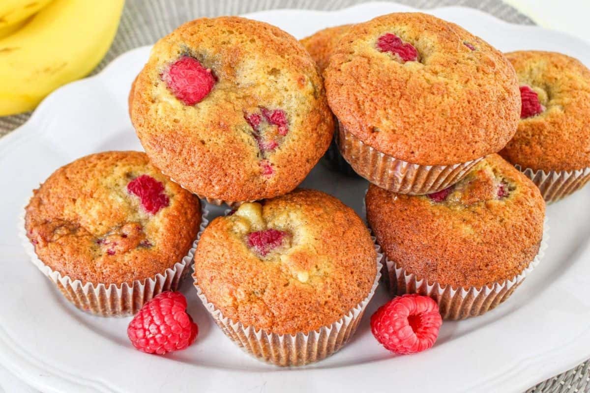 A white platter stacked with banana raspberry muffins with whole raspberries and bananas in the background.