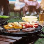 Assorted vegetables and steaks grilling on a barbecue, nodding to the history of American BBQ, with corn on the cob and a bottle of beer nearby; people sit at a picnic table in the background.