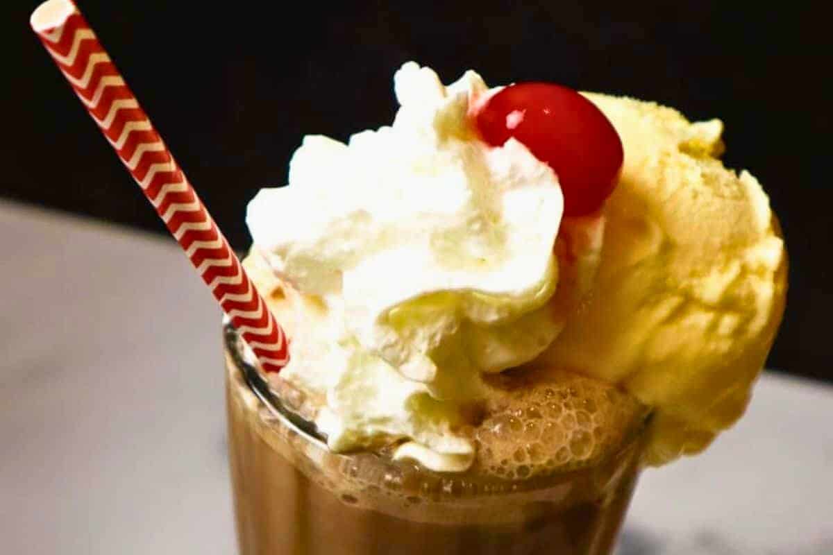 The image shows a milkshake with a red and white straw, topped with vanilla ice cream, whipped cream, and a cherry.