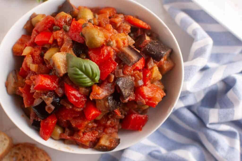 A bowl of Caponata with diced eggplant, red bell peppers, tomatoes, onions, and a basil garnish. A blue and white striped cloth is beside the bowl.