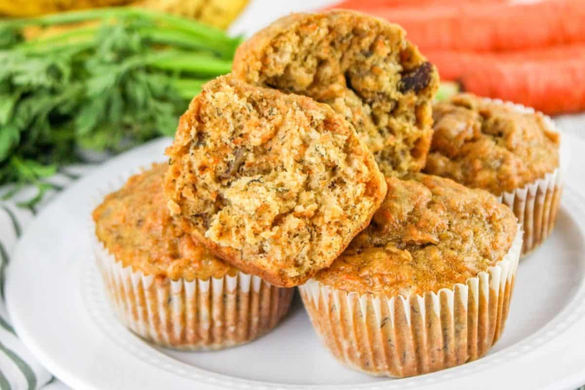 A stack of carrot banana muffins on a white plate, surrounded by fresh carrots and a striped cloth.