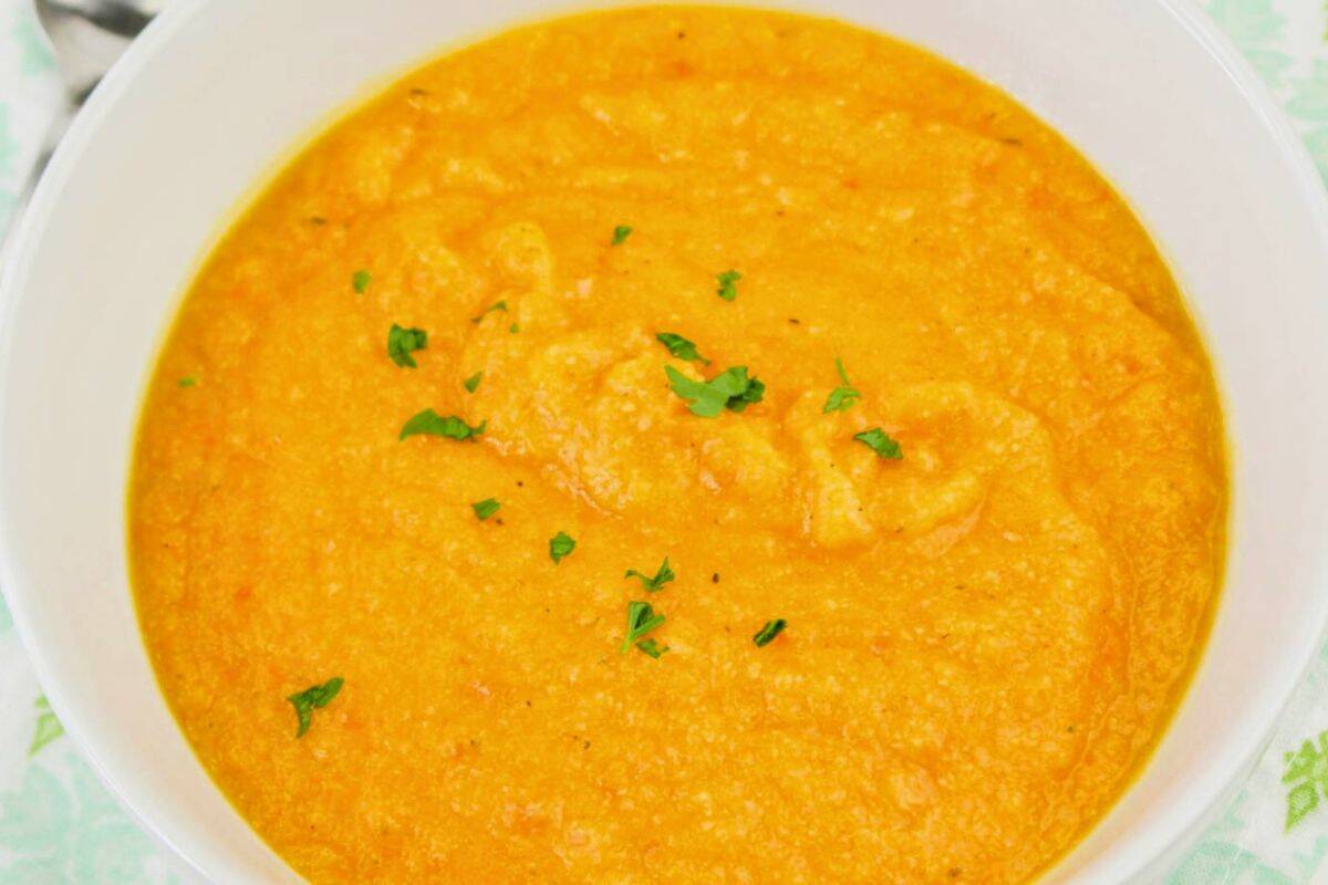 Bowl of creamy carrot soup garnished with small parsley pieces, placed on a patterned green and white napkin.