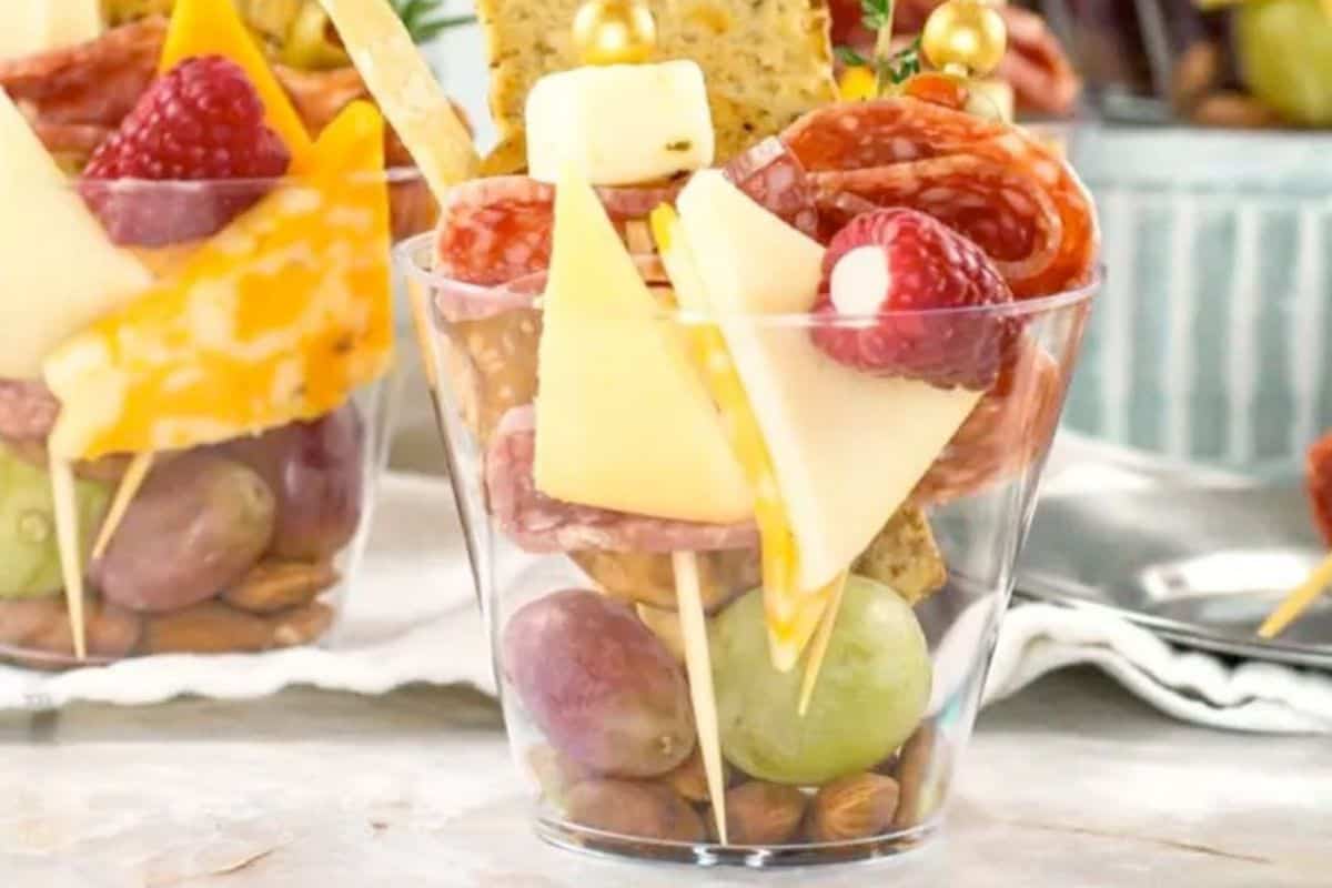 A clear plastic cup filled with grapes, almonds, cheese cubes, salami slices, a raspberry, and crackers, arranged on skewers for a single-serve charcuterie snack.