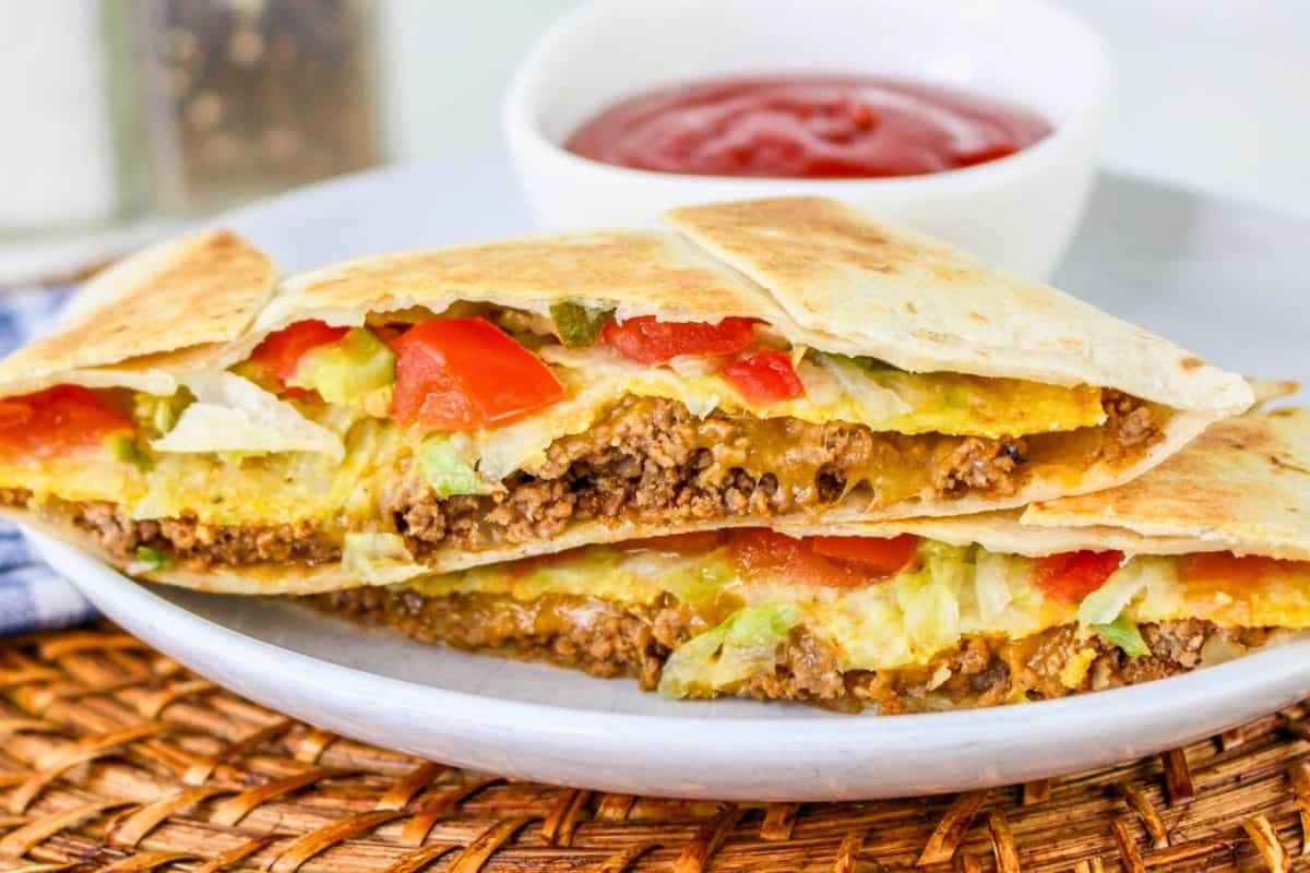 A sliced quesadilla filled with seasoned ground beef, cheese, lettuce, and tomatoes on a plate, accompanied by a small bowl of red sauce.