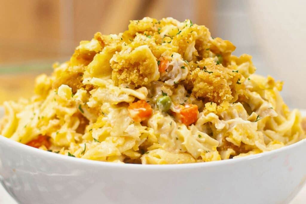 A white bowl filled with creamy pasta mixed with breaded chicken pieces, peas, carrots, and a sprinkling of herbs.