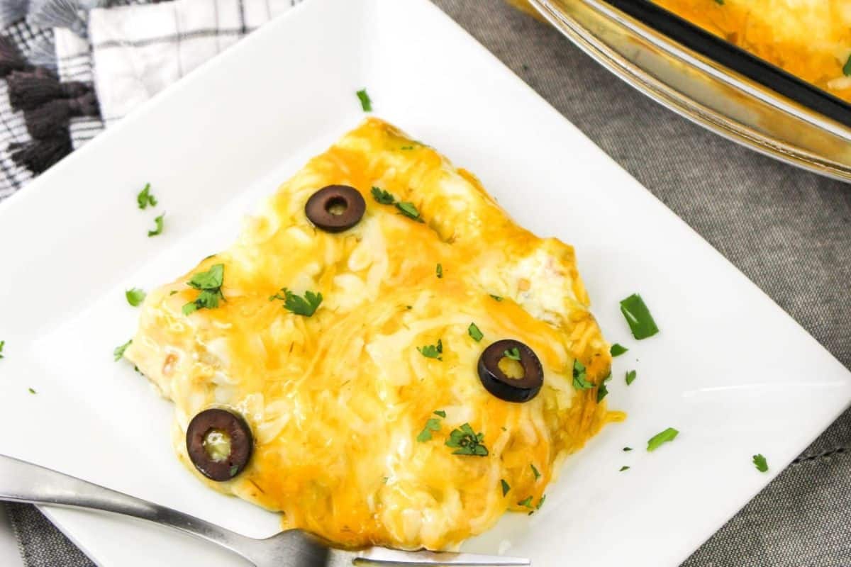 A plate of chicken taquito topped with melted cheese, black olive slices, and chopped parsley. A fork is placed on the plate, and a dish of olives is partially visible in the background.