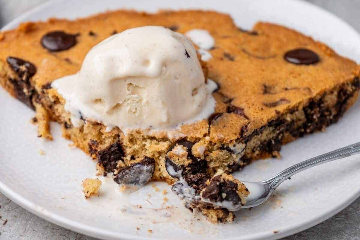 A slice of chocolate chip skillet cookie topped with a scoop of vanilla ice cream sits on a white plate with a spoon.