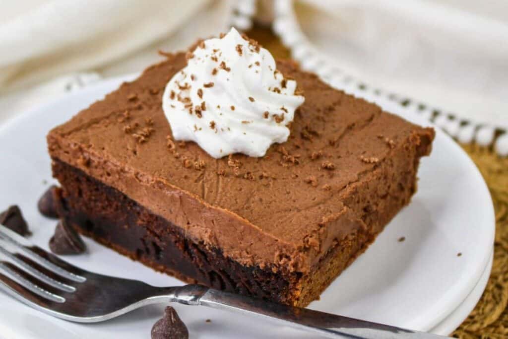 A slice of chocolate mousse cake topped with whipped cream and chocolate shavings on a white plate, next to a fork and chocolate chips.
