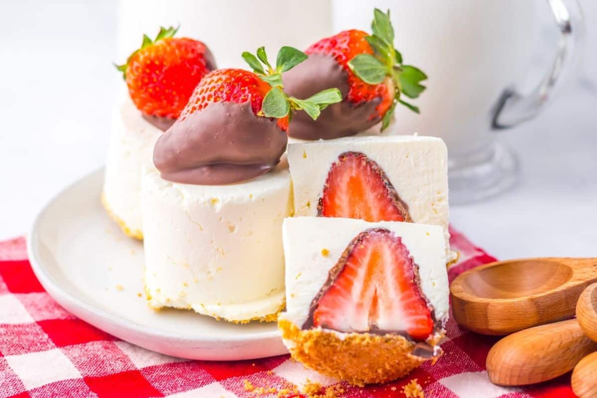 Cheesecake slices topped with chocolate-covered strawberries on a white plate. Red checkered cloth and wooden spoons beside.