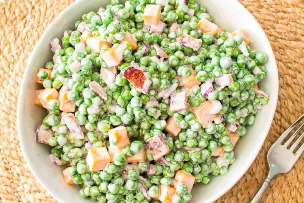 A bowl of pea salad with diced cheese, bacon pieces, and creamy dressing on a woven placemat, next to a fork.