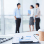 A paper cup, laptop, and documents with charts sit on a desk; three people stand and talk in the blurred background near large windows, perhaps discussing plans to leave the office early.