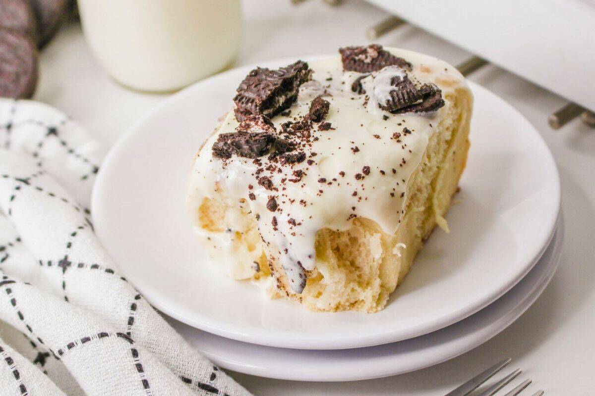 A cinnamon roll topped with white icing and crushed chocolate cookies on a white plate, next to a napkin, a fork, and milk.