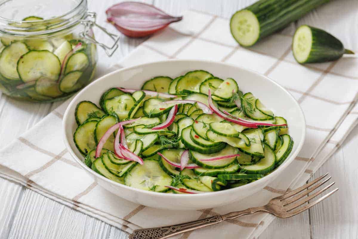 A white bowl filled with Quick Marinated Cucumbers sits on a checked cloth next to a fork, with a glass jar of more salad and whole cucumbers in the background.