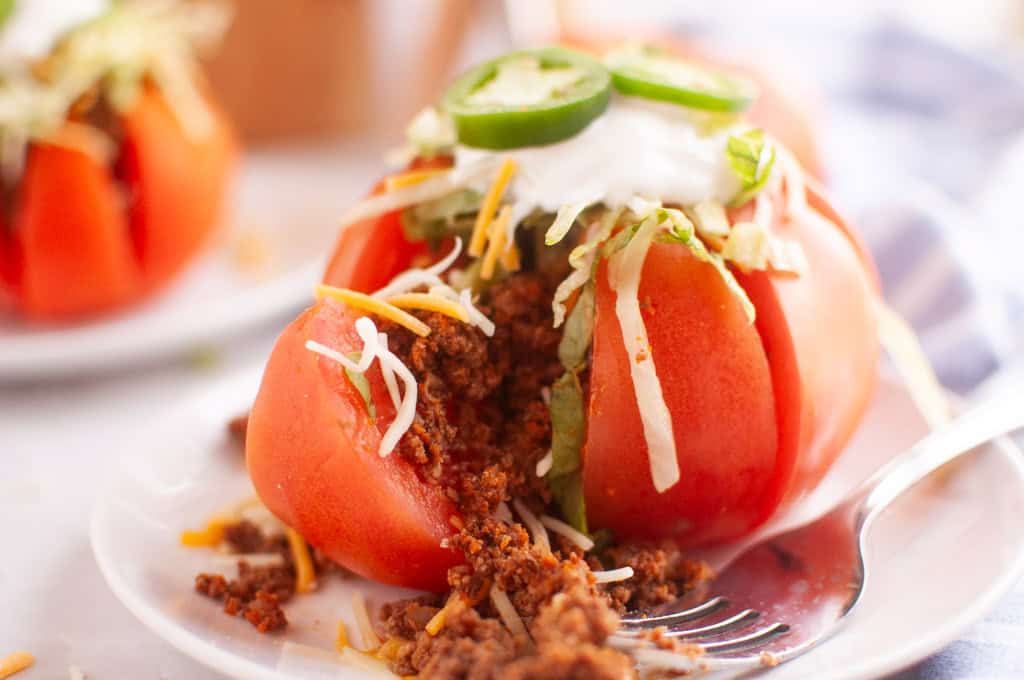 A sliced tomato stuffed with seasoned ground meat, shredded cheese, lettuce, sour cream, and jalape&ntilde;o slices is served on a white plate with a fork.