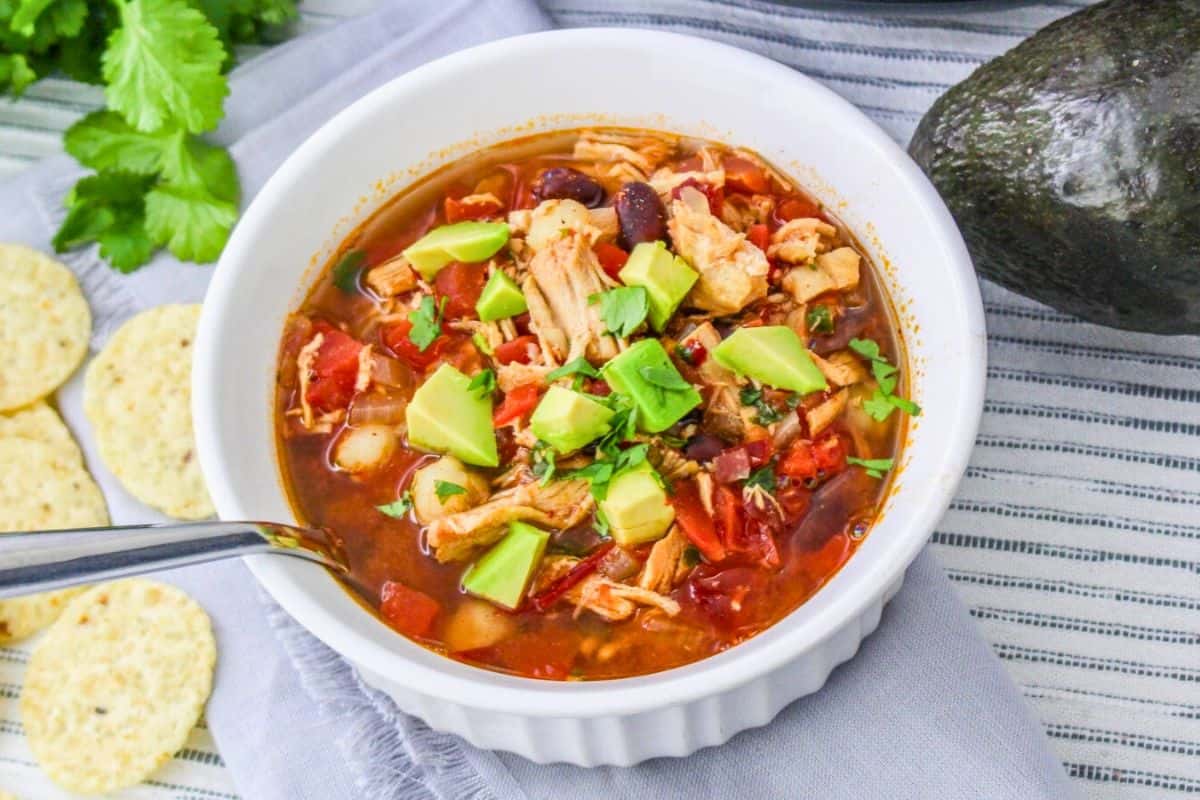 Bowl of chicken soup with avocado and cilantro on top, spoon in bowl, tortilla chips and avocado beside, placed on a striped cloth.