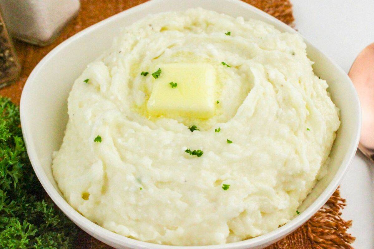 Bowl of mashed potatoes with a pat of butter on top, garnished with parsley. A spoon and a sprig of parsley are next to the bowl.