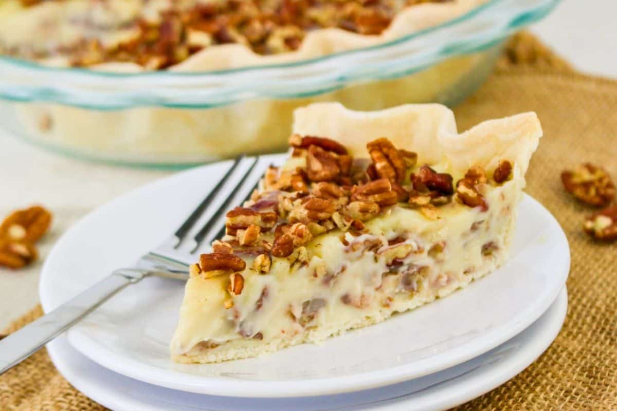 Slice of pecan pie on a white plate with a fork, placed on a burlap surface. The pie has a pastry crust, creamy filling, and is topped with chopped pecans.