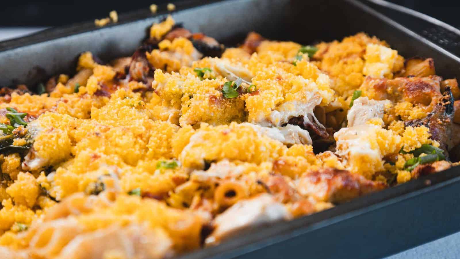 A close-up of a baked pasta dish in a rectangular metal pan with a golden breadcrumb topping, pieces of pasta, baked cheese, and green herbs visible.