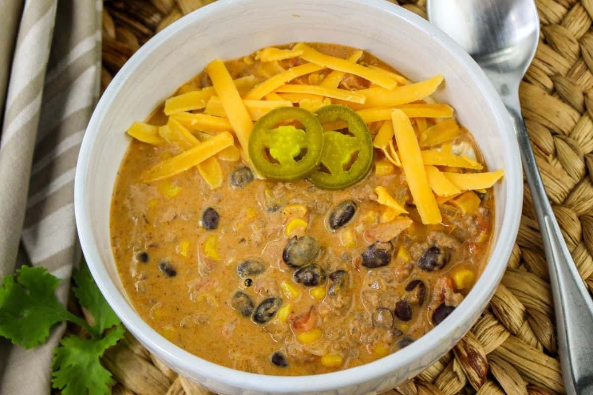 A bowl of creamy soup with black beans, corn, shredded cheese, and jalapeño slices, placed on a woven mat next to a spoon and cloth napkin.