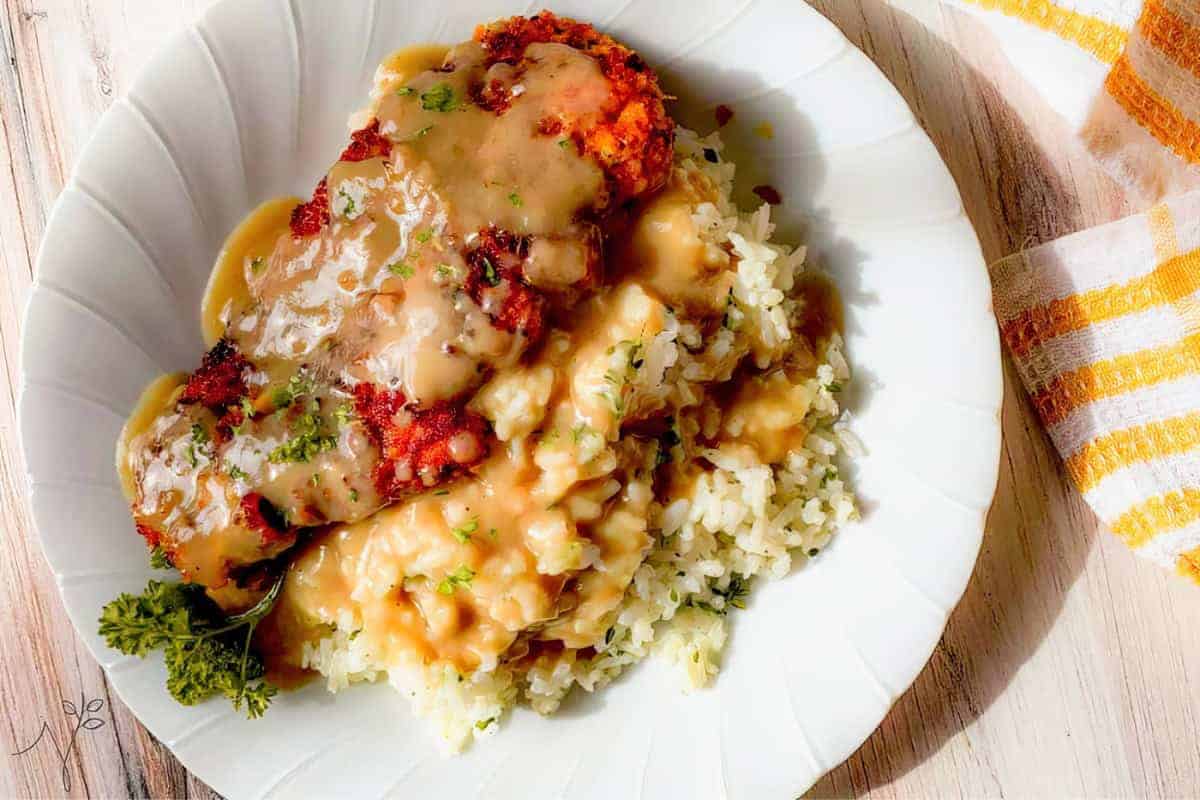 A plate of breaded and fried chicken cutlet topped with brown gravy sits on a bed of white rice. The dish is garnished with chopped fresh herbs and accompanied by a sprig of parsley. A striped yellow and white napkin is placed beside the plate on a wooden table.