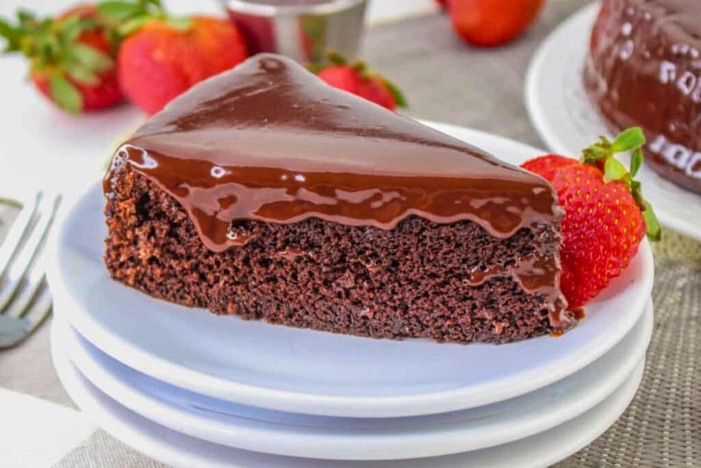 A slice of chocolate fudge cake with glossy chocolate icing on a white plate, garnished with a fresh strawberry; whole strawberries in the background.