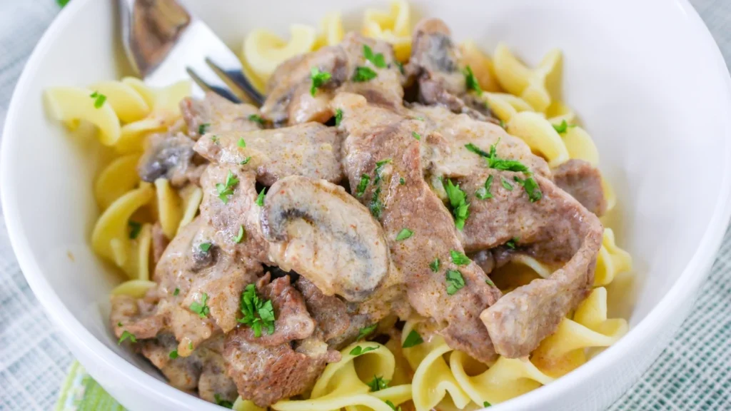 A white bowl filled with egg noodles topped with beef stroganoff, including sliced beef, mushrooms, creamy sauce, and chopped parsley. This comforting one-pot recipe is served with a fork resting in the bowl.