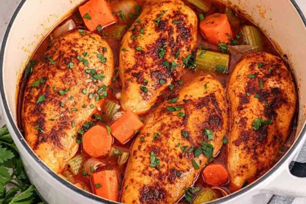 Four seasoned chicken breasts simmer in a pot with chopped carrots, celery, onions, and herbs in a savory broth.