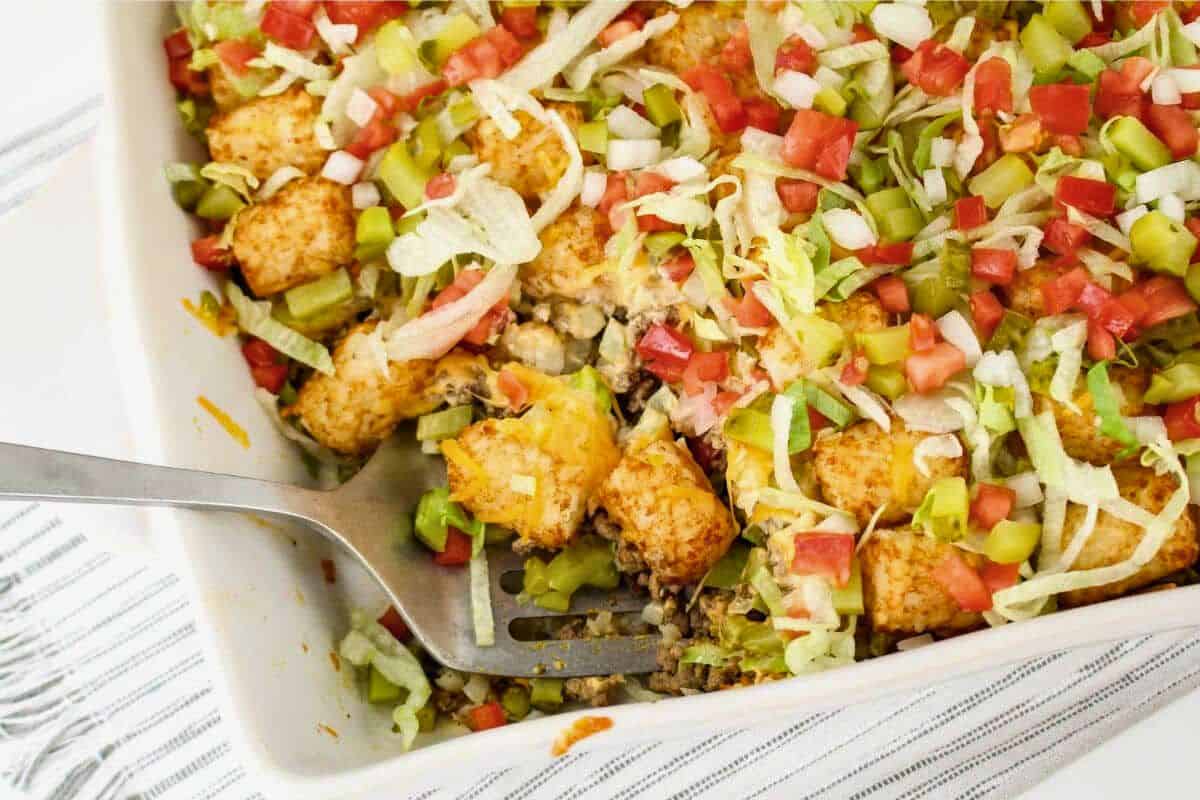 A casserole dish filled with tater tots, ground beef, diced tomatoes, shredded lettuce, onions, and pickles, with a serving spoon on the side.